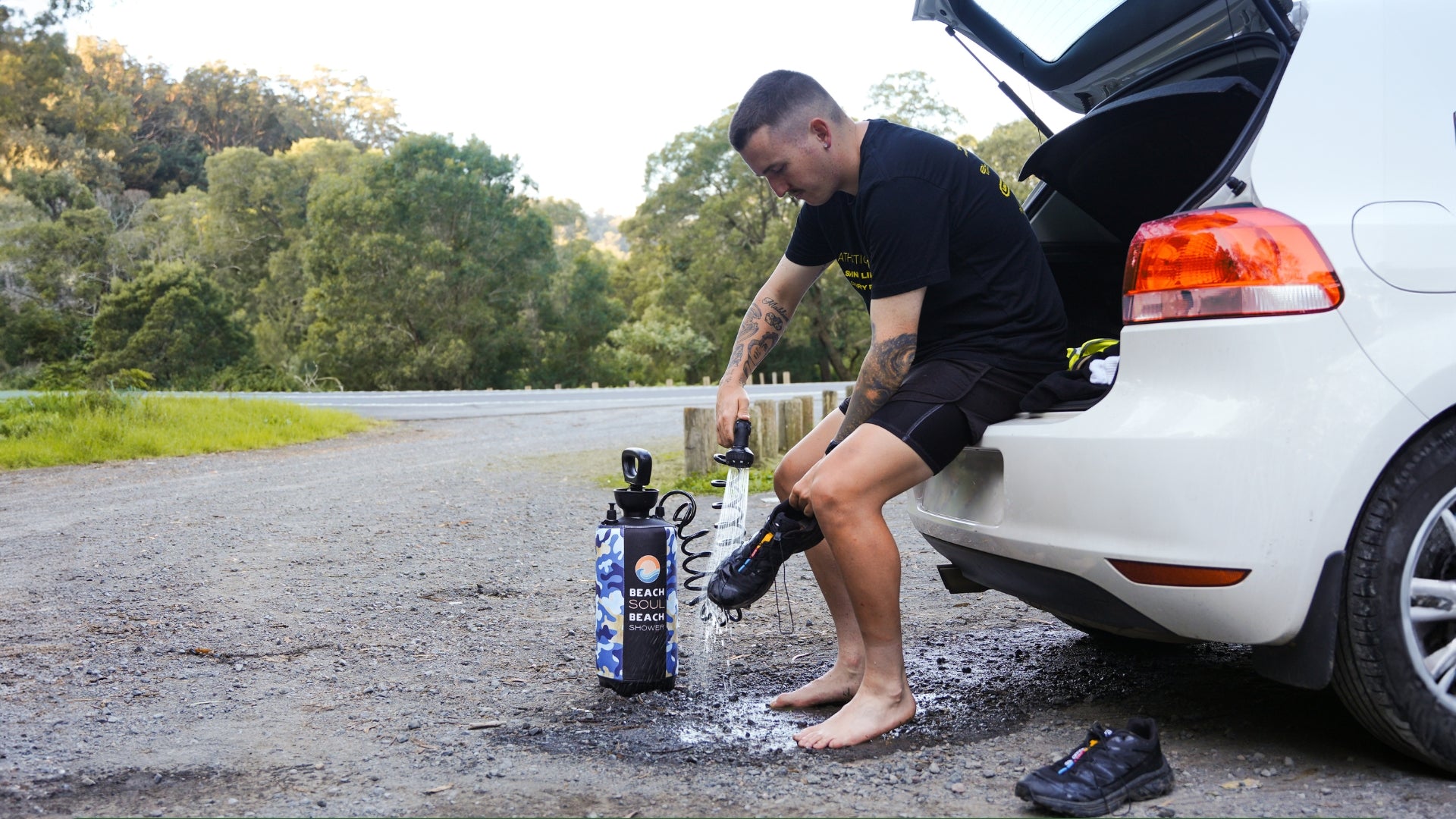 Man rinsing an outdoor shoe beside a car with a Beach Soul portable shower