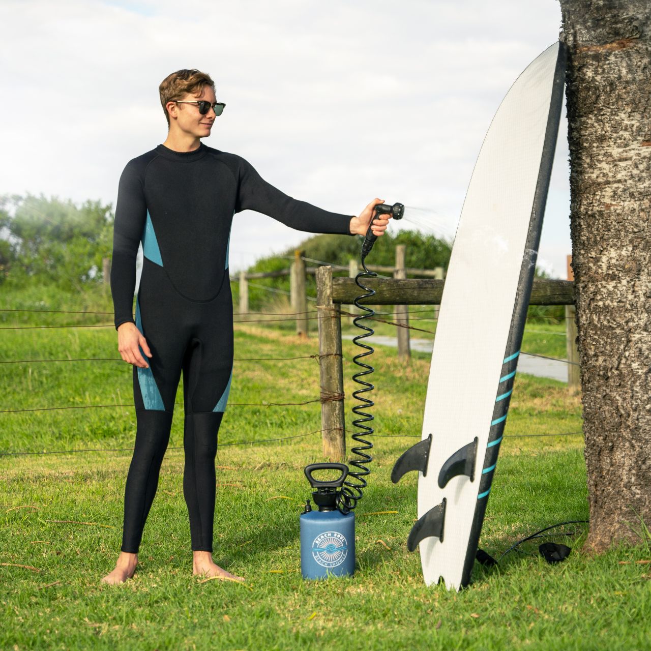 Surfer rinsing sand of his surfboard with the Ocean Rise portable shower on the grass