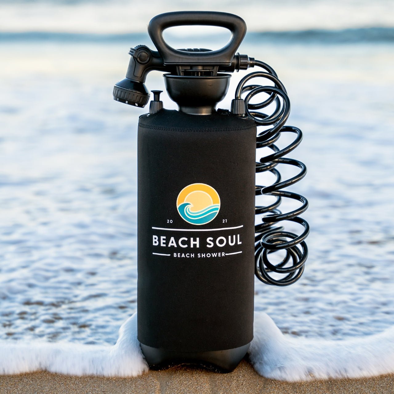 A black portable shower with a 'BEACH SOUL' logo, manual pump, and coiled hose, placed on the sand with the ocean in the background.