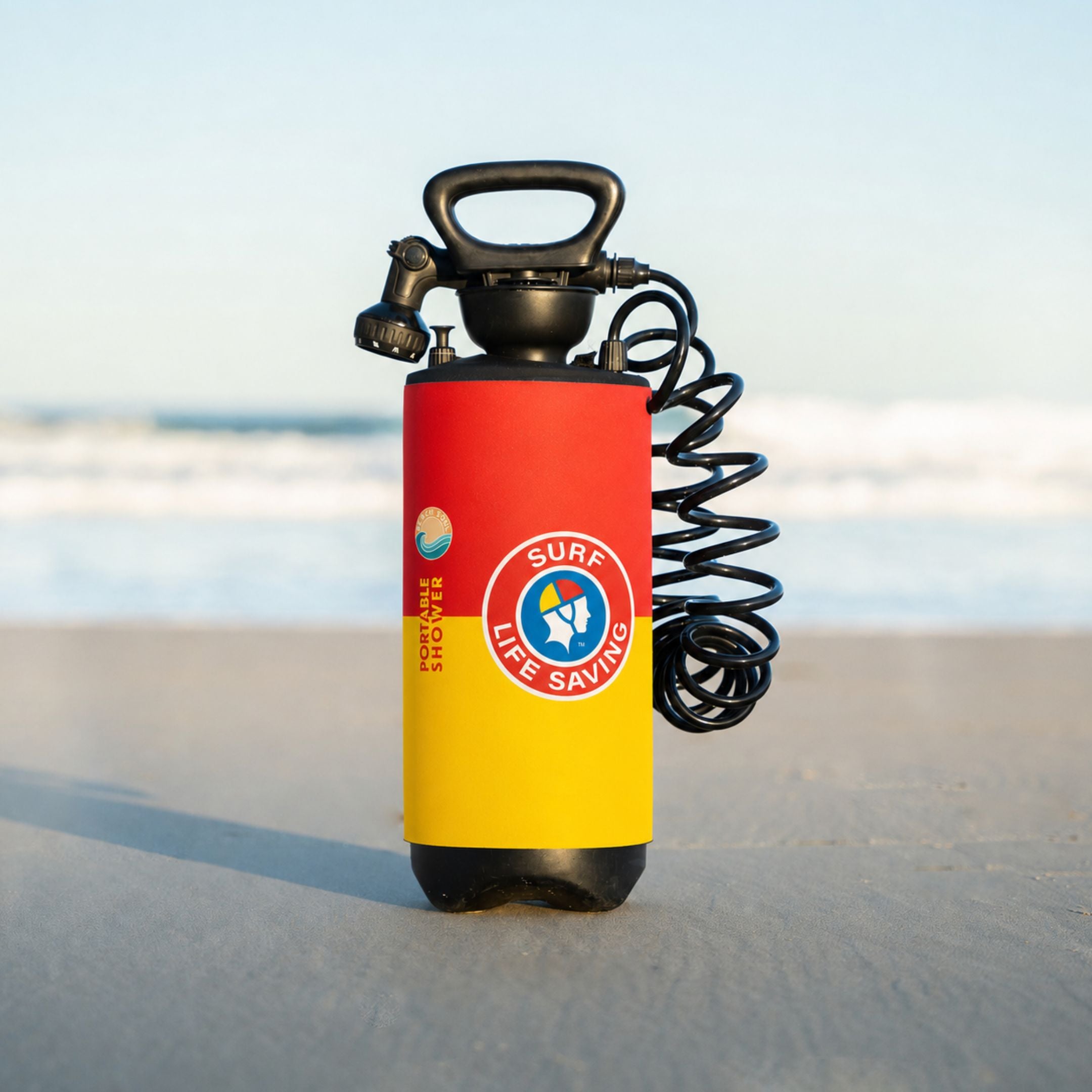 A Surf Life Saving branded portable shower tank with a black hose, standing on a sandy beach with the ocean in the background.