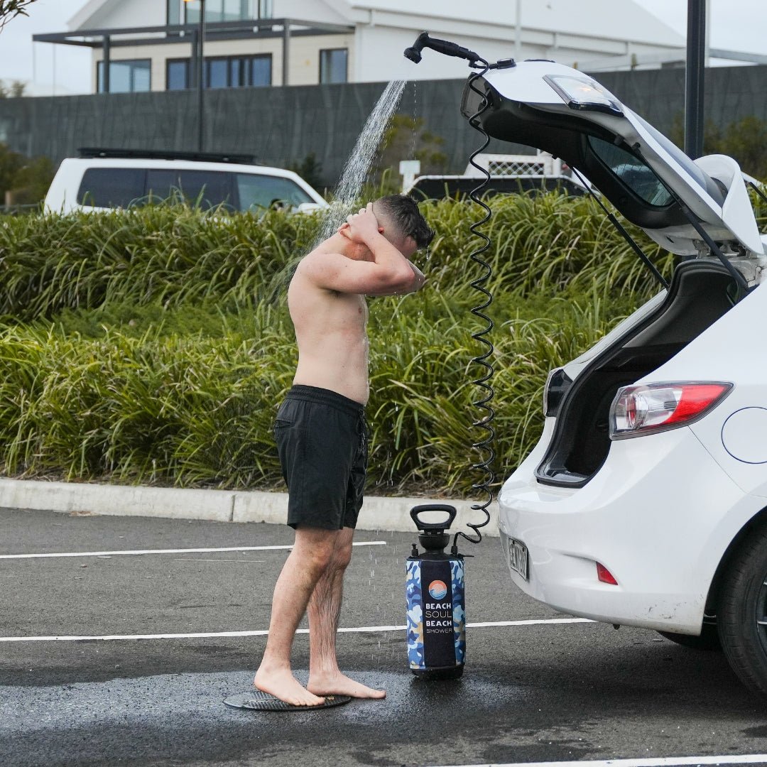 Man showering with a Beach Soul portable shower while standing on the rinse mat from the Bag 'n' Mat beside a car trunk