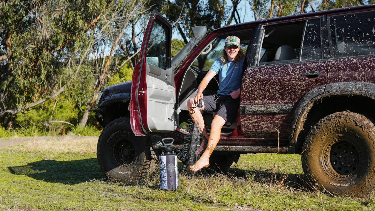 Woman rinsing muddy legs with a Beach Soul portable shower while seated in a 4WD after an off-road adventure