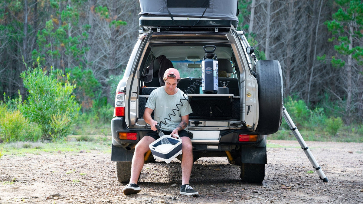 Man washing outdoor gear with a Beach Soul portable shower while sitting on the back of a 4WD at a remote bush campsite