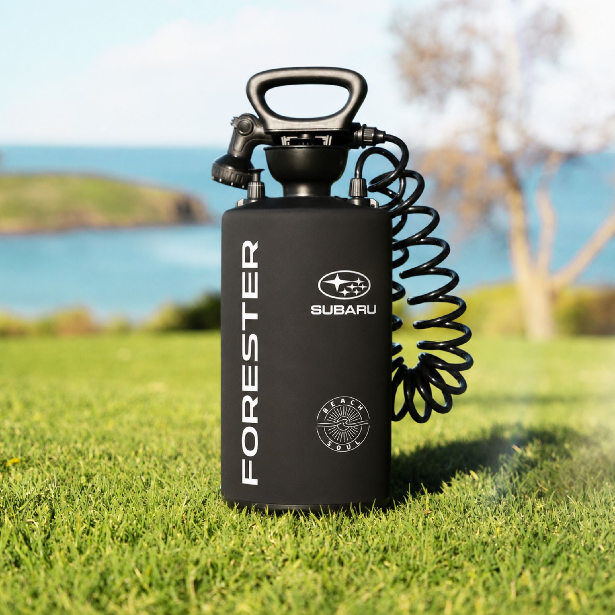 A black Forester Subaru brand pressurized shower with a coiled hose, standing on a green grass lawn with a blurred background of blue water and sky.
